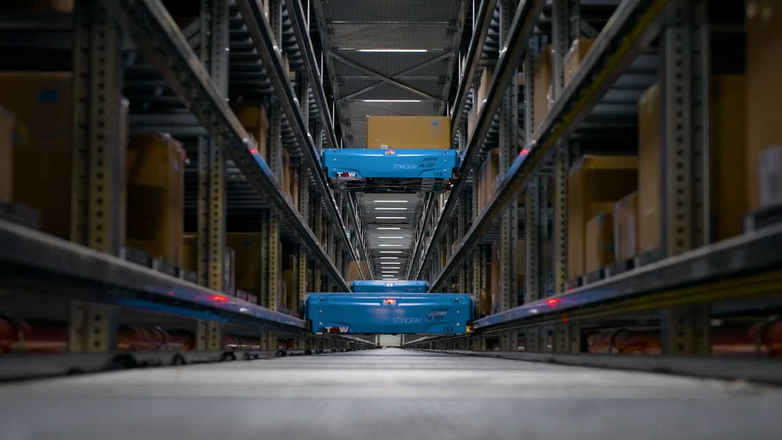 Two TGW Logistics Stingray shuttles with blue covers in an aisle using trays for storage