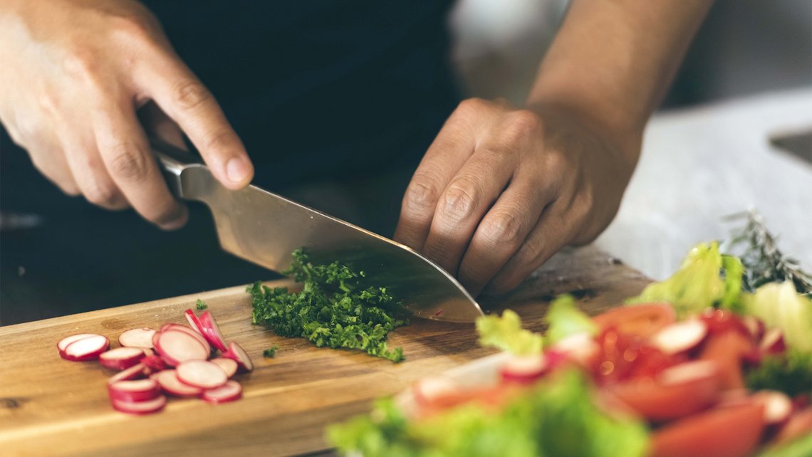 [Translate to France:] Hands of a man cutting vegetables and herbs on a wooden board