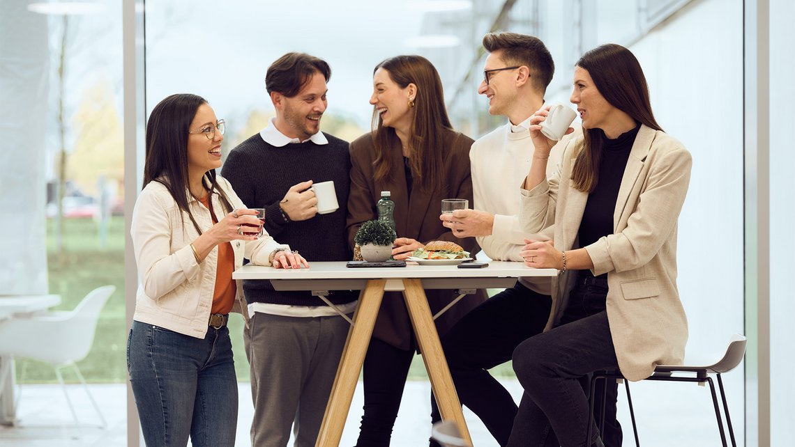 Five TGW Logistics employees laughing at a standing table during a team coffee break