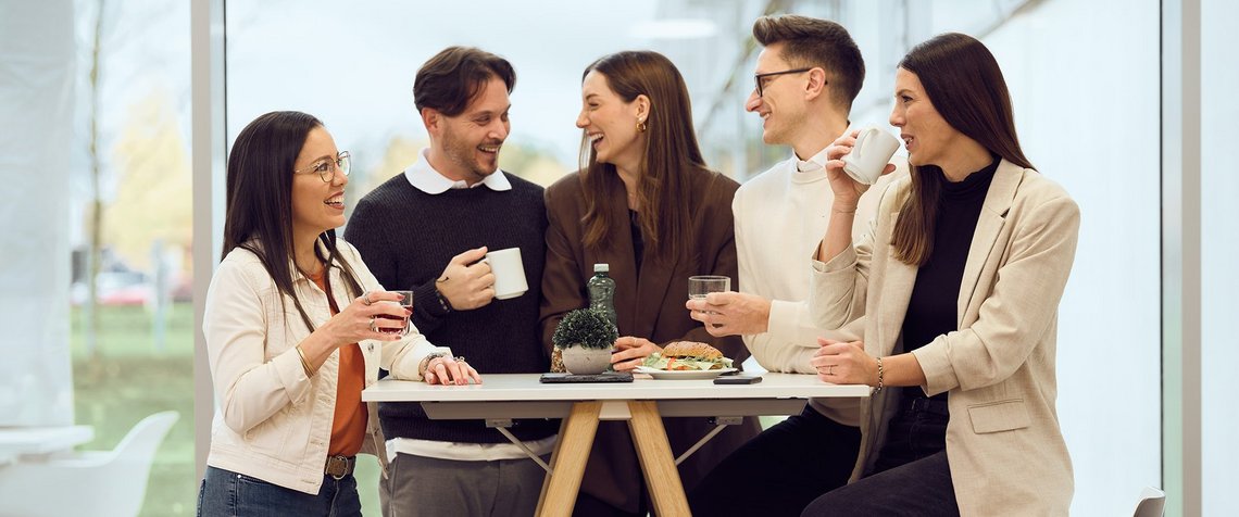 Five TGW Logistics employees laughing at a standing table during a team coffee break