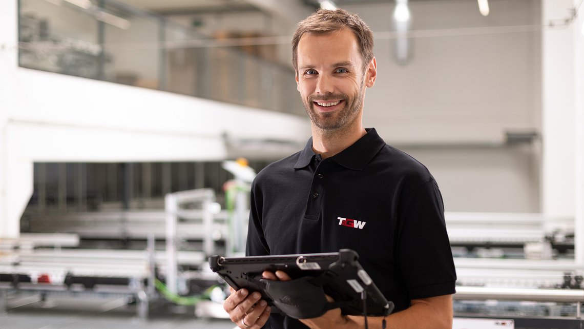 [Translate to France:] A TGW service employee smiles into the camera, holding a tablet in his hand