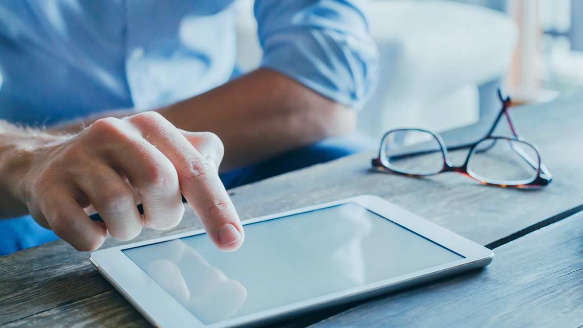 Close up of a hand scrolling through news on their tablet, which is resting on the table next to their glasses