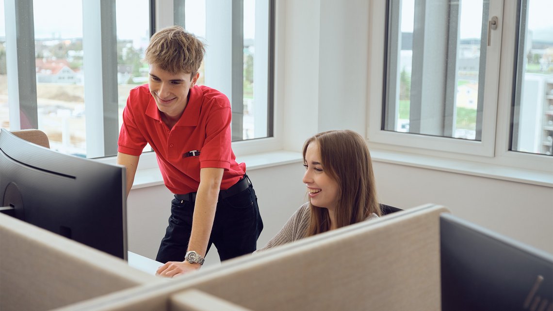 A TGW Logistics apprentice looking at a desktop screen while receiving information from a TGW employee at a desk